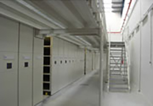 Pasillo interior de depósito con armarios compactos, estructura metálica y escalera bajo lucernario.