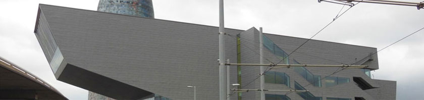 Edificio gris de volúmenes angulares con ventanales; cables de tranvía en primer plano y cielo nublado.
